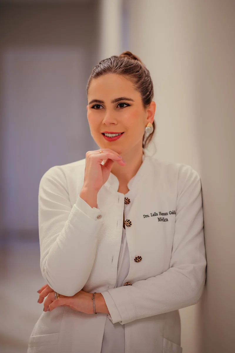 Dra. Laila Hassan sorrindo, de jaleco branco, com uma mão próxima ao rosto.
